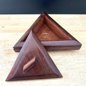 May include: A triangular wooden jewelry box with a hinged lid. The box is open, revealing two gold rings inside. The box is made of dark brown wood and sits on a light-colored wooden surface.