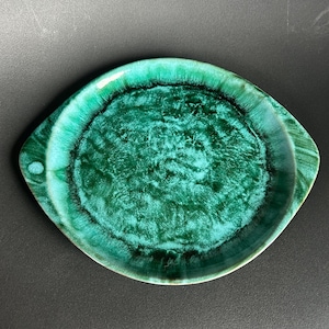 May include: A green ceramic serving dish with a unique, eye-shaped design. The dish features a glossy, textured surface with shades of green and a dark green center. It has two small handles on either side.