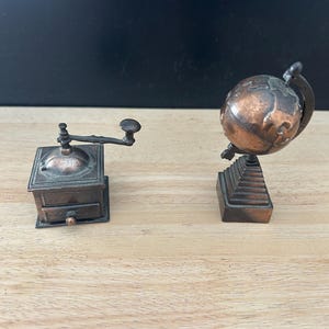 Puede incluir: Dos objetos decorativos en miniatura de color cobre: un molinillo de café con manivela y un globo terráqueo sobre una base escalonada. Los objetos están colocados sobre una superficie de madera clara con un fondo oscuro.