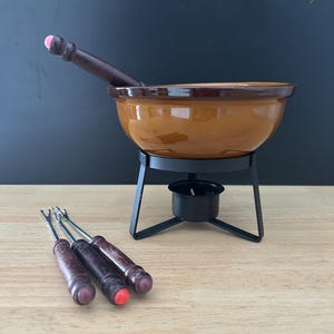 Puede incluir: Un juego de fondue vintage con una olla de cerámica marrón, un mango de madera oscura y un soporte de metal negro. Tres tenedores de fondue con mangos de madera y puntas de colores están en primer plano. El juego está diseñado para derretir queso o chocolate.