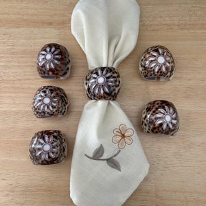 Könnte beinhalten: Ein Set aus sechs dekorativen Serviettenringen mit braunem und weißem Sprenkelmuster, jeweils mit einem weißen Blumendesign. Eine cremefarbene Serviette mit floraler Stickerei befindet sich in der Mitte. Die Artikel sind auf einer hellbraunen Holzoberfläche angeordnet.