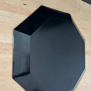 May include: A black, geometric-shaped plate with a glossy finish. The plate has an octagonal shape with beveled edges. The plate is sitting on a light-colored wooden surface.