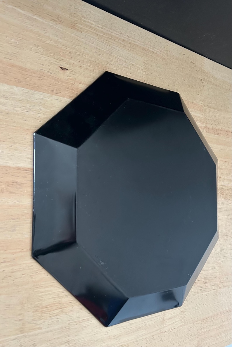 May include: A black, geometric-shaped plate with a glossy finish. The plate has an octagonal shape with beveled edges. The plate is sitting on a light-colored wooden surface.