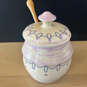 May include: A ceramic honey pot with a light beige base, featuring hand-painted lavender bands and dark blue scalloped line patterns. The pot includes a matching lid with a knob handle and a light-coloured wooden honey dipper resting inside.