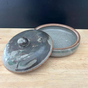 Puede incluir: Un recipiente de cerámica hecho a mano con tapa. El recipiente es de color azul-verdoso apagado con ribete marrón. La tapa tiene un pequeño pomo y un patrón moteado de gris oscuro y verde. El recipiente está sobre una superficie de madera clara.
