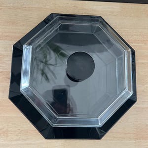 May include: An octagonal black and clear plastic container with a black knob on the lid. The container has a glossy finish and is sitting on a light brown wooden surface.