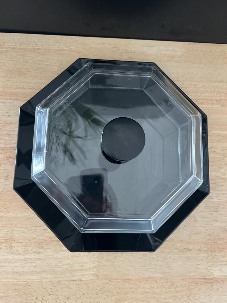 May include: An octagonal black and clear plastic container with a black knob on the lid. The container has a glossy finish and is sitting on a light brown wooden surface.