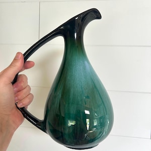 Puede incluir: Una jarra de cerámica en forma de lágrima con un acabado brillante y un esquema de color degradado que transita desde un verde bosque profundo y oscuro en la base y el asa hasta un tono cerceta más claro y vibrante cerca del pico vertedor.
