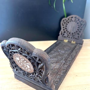 May include: Dark brown wooden book stand with intricate floral carvings. The stand features a decorative oval inlay with white floral designs. The back of the stand is also carved with a floral pattern. The stand is hinged and can be folded flat.