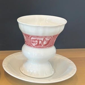 May include: A white porcelain egg cup with a flared rim and a pedestal base, resting on a matching circular saucer. The cup features a horizontal band decorated with a red printed landscape scene enclosed in geometric frames around the centre.