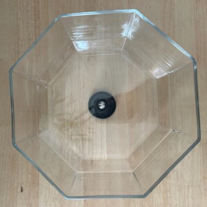 May include: A clear, octagonal glass bowl with a black, circular object in the center. The bowl is empty and sits on a light brown wooden surface. The bowl's edges are clearly defined.