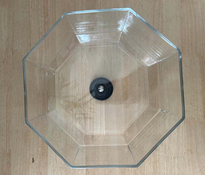 May include: A clear, octagonal glass bowl with a black, circular object in the center. The bowl is empty and sits on a light brown wooden surface. The bowl's edges are clearly defined.