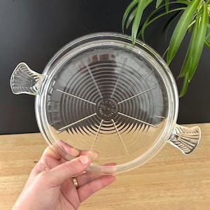 May include: Clear glass Pyrex lid with two handles. The lid has a circular design with concentric rings and radial lines. The text "PYREX" and "MADE IN USA" are visible. The lid is designed for cookware.