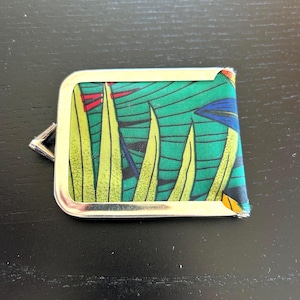 May include: A rectangular, silver-framed coin purse with a colourful fabric insert. The fabric features a green background with yellow and blue plant-like designs. The purse has a small, silver clasp.