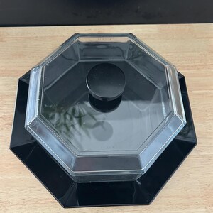 May include: A black, octagonal serving dish with a clear, octagonal lid and a black circular handle. The dish sits on a wooden surface with a black background.