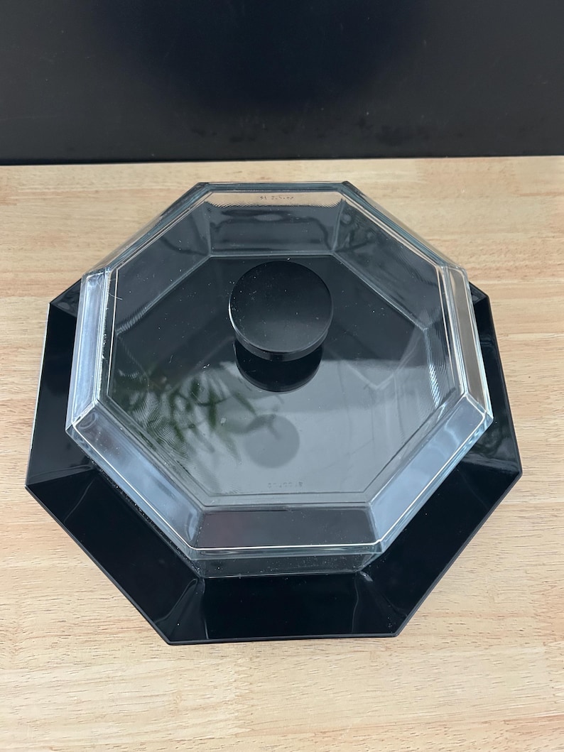 May include: A black, octagonal serving dish with a clear, octagonal lid and a black circular handle. The dish sits on a wooden surface with a black background.