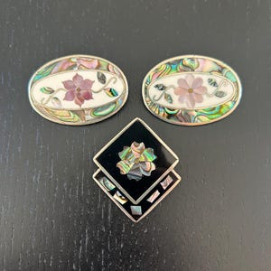 May include: Three decorative brooches with floral designs. Two oval brooches feature a white background with a pink flower and abalone shell accents. The third brooch is square with a black background and a flower design.