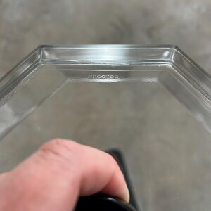 May include: Clear glass hexagonal container with the word "arcofoc" etched on the side. The container is held by a hand, with a black object visible at the bottom. The background is a blurred gray.