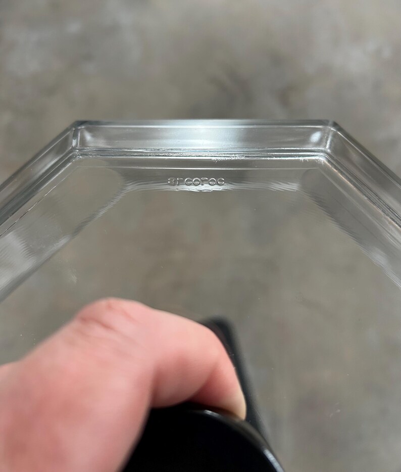 May include: Clear glass hexagonal container with the word "arcofoc" etched on the side. The container is held by a hand, with a black object visible at the bottom. The background is a blurred gray.