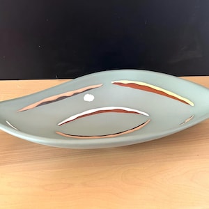 May include: An elongated, leaf-shaped ceramic bowl in a matte sage green finish. The interior surface features a series of curved, abstract decorative accents in shades of terracotta, cream, and dark brown, arranged in a rhythmic, flowing pattern.
