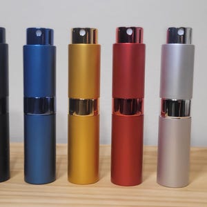 May include: Five perfume atomisers in black, blue, gold, red, and silver. Each has a cylindrical body and a spray nozzle. The atomisers are arranged in a row on a wooden surface.