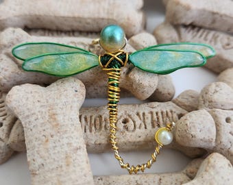 May include: A handcrafted dragonfly brooch with a teal and gold head, green and yellow wings, and a gold and green wire body. The brooch has a pearl accent and is set against a background of dog biscuits.