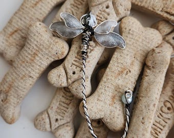 May include: A handcrafted dragonfly brooch with silver wings and a black and silver wire body. The brooch is set against a background of dog biscuits, creating a whimsical contrast. The dragonfly's tail ends with a small, light-colored bead.