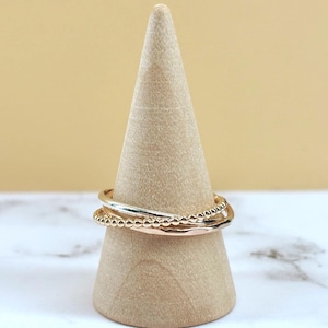 Puede incluir: Un conjunto de tres anillos apilados sobre un cono de madera. El set incluye una banda dorada lisa, una banda dorada con textura de cuentas y una banda de oro rosa lisa, todas con un acabado metálico pulido y perfiles delgados.