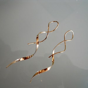 May include: A pair of gold-colored, spiral-shaped earrings. The earrings have a long, curved design with a loop at the top and a tapered end. The earrings are displayed on a reflective surface.