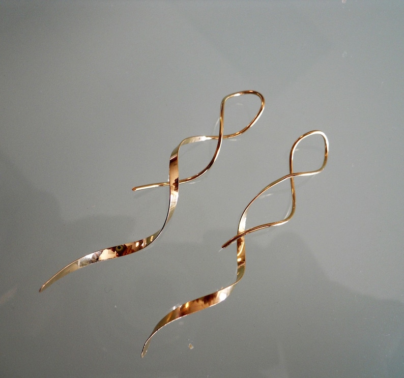May include: A pair of gold-colored, spiral-shaped earrings. The earrings have a long, curved design with a loop at the top and a tapered end. The earrings are displayed on a reflective surface.