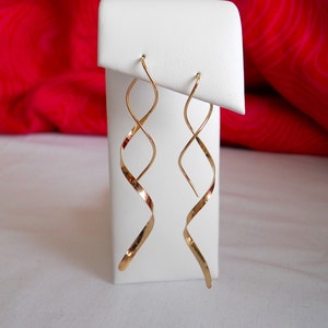 May include: A pair of gold-colored, spiral-shaped earrings. The earrings are displayed on a white stand against a red background. The earrings have a long, slender design with a twisted, ribbon-like appearance.