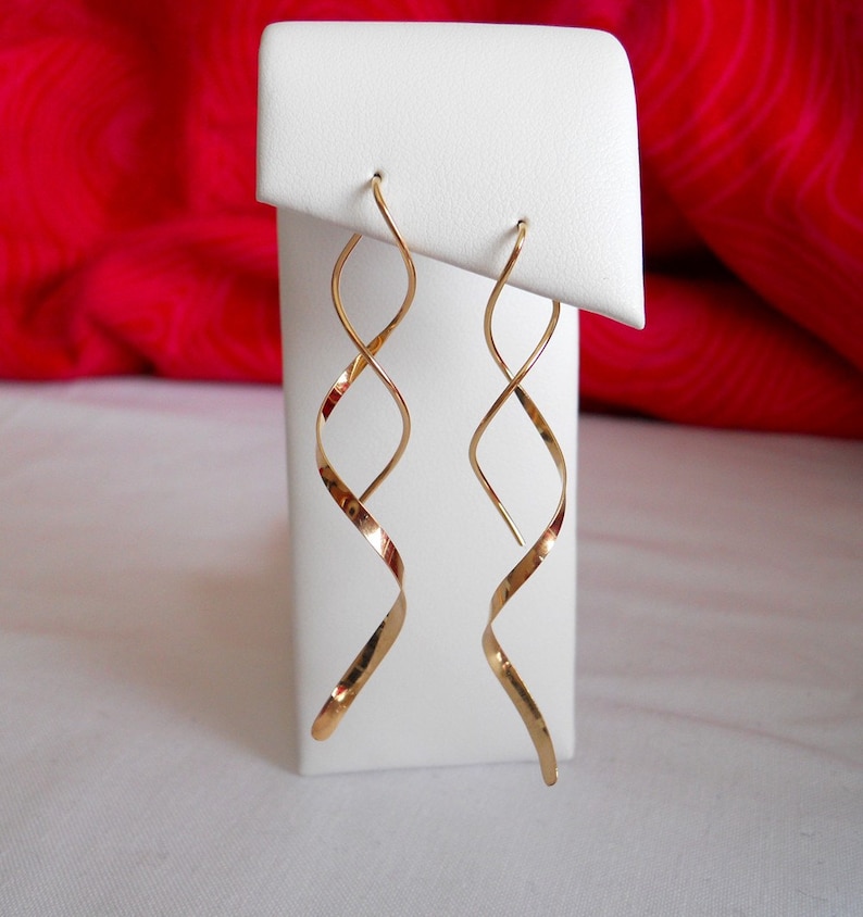 May include: A pair of gold-colored, spiral-shaped earrings. The earrings are displayed on a white stand against a red background. The earrings have a long, slender design with a twisted, ribbon-like appearance.