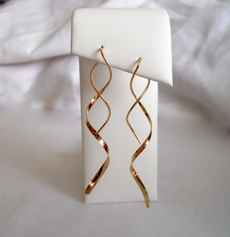May include: A pair of gold-colored spiral earrings. The earrings are displayed on a white stand against a white background. The earrings have a long, flowing design, with a hook at the top for the ear.