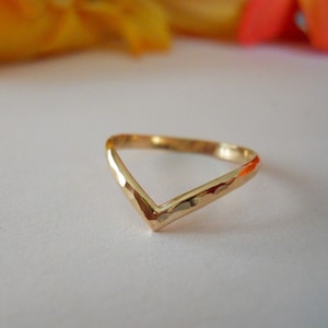 Puede incluir: Un anillo dorado en forma de V con una textura martillada en la superficie exterior. La banda presenta un diseño de chevrón puntiagudo que crea una forma angular distintiva, con un acabado pulido que refleja la luz sobre su superficie.
