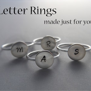 Puede incluir: Cuatro anillos de plata con tapas redondas y planas, cada uno grabado con una letra diferente: M, R, A y S. Los anillos están dispuestos en semicírculo sobre una superficie gris. Anillos de letras hechos solo para ti.