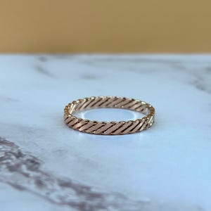 May include: A rose gold ring featuring a continuous twisted rope or braided metal design. The band has a uniform width and a polished, reflective metallic finish that catches the light, creating a series of diagonal grooves around the entire circumference.