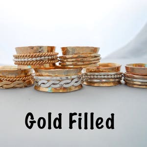 May include: Five gold-filled spinning rings with different designs. The rings are stacked on a white surface. The text "Gold Filled" is written below the rings.