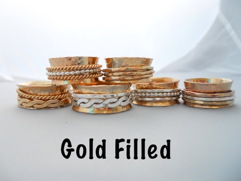 May include: Five gold-filled spinning rings with different designs. The rings are stacked on a white surface. The text "Gold Filled" is written below the rings.
