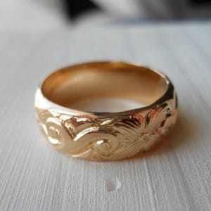 Puede incluir: Un anillo de banda de oro pulido con un intrincado patrón en relieve de volutas y un motivo floral central. La superficie metálica refleja la luz, resaltando los grabados detallados que envuelven toda la circunferencia del anillo.