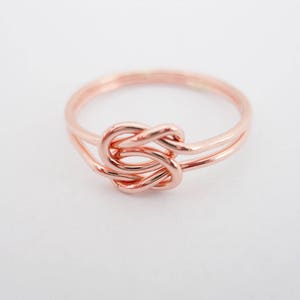 Puede incluir: Un anillo de nudo de oro rosa con un diseño simple. El anillo está hecho de dos bandas de metal entrelazadas.