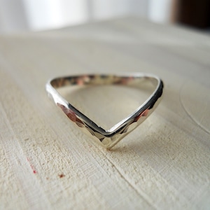 Könnte beinhalten: Ein silberfarbener V-förmiger Ring mit einer gehämmerten Textur entlang des äußeren Bandes. Die Metalloberfläche hat ein reflektierendes Finish mit ausgeprägten Vertiefungen, die durch den Hammervorgang entstanden sind und eine spitze Chevron-Silhouette bilden.