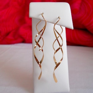 May include: A pair of gold-toned dangle earrings with a twisted, spiral design. The earrings are displayed on a white stand.