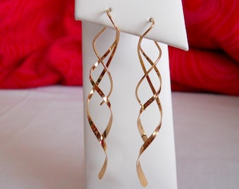 May include: A pair of gold-toned dangle earrings with a twisted, spiral design. The earrings are displayed on a white stand.