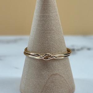 May include: A thin gold-coloured ring featuring a delicate twisted knot design at the centre. The band is smooth and polished, showcasing a continuous metallic finish that wraps around the finger. The ring is displayed on a wooden cone stand.