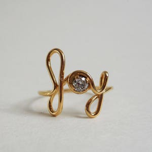 May include: Gold ring with a diamond set in the center, spelling out the word "Joy" in a cursive font.