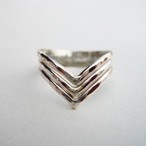 May include: A silver-toned chevron ring featuring three stacked, V-shaped bands. The metal surface has a hammered, textured finish that catches the light, creating a layered appearance with a pointed center design that forms a distinct V shape.