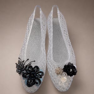 May include: A pair of clear, jelly-style flats with a textured, woven-look exterior and a silver-toned interior sole. Each shoe features a decorative cluster of black and dark blue floral-shaped embellishments, beads, and sequins on the toe.