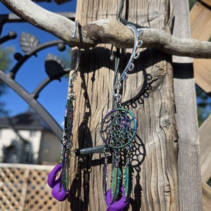 Puede incluir: Un par de pendientes hechos a mano con un diseño de atrapasueños. Los pendientes tienen un acabado metálico plateado con un atrapasueños circular, detalles morados y plumas colgantes. Los pendientes están suspendidos de una rama de madera.