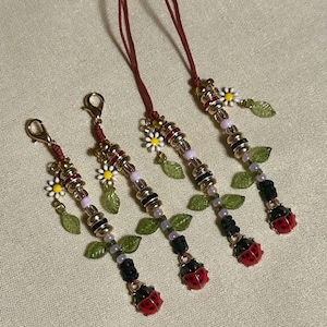 May include: Four beaded ladybug charms with gold-colored clasps and red cords. Each charm features a red ladybug, green leaf accents, white daisy, and various beads in gold, black, and pink. The charms are arranged on a beige surface.