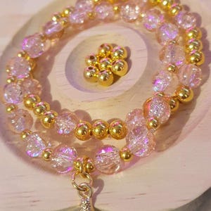 Op de afbeelding: Een armband met een gouden zeemeerminstaartbedel. De armband heeft iriserende roze en heldere kralen afgewisseld met goudkleurige kralen. Een kleine hoop gouden kralen ligt in de buurt. De armband wordt op een houten oppervlak gepresenteerd.
